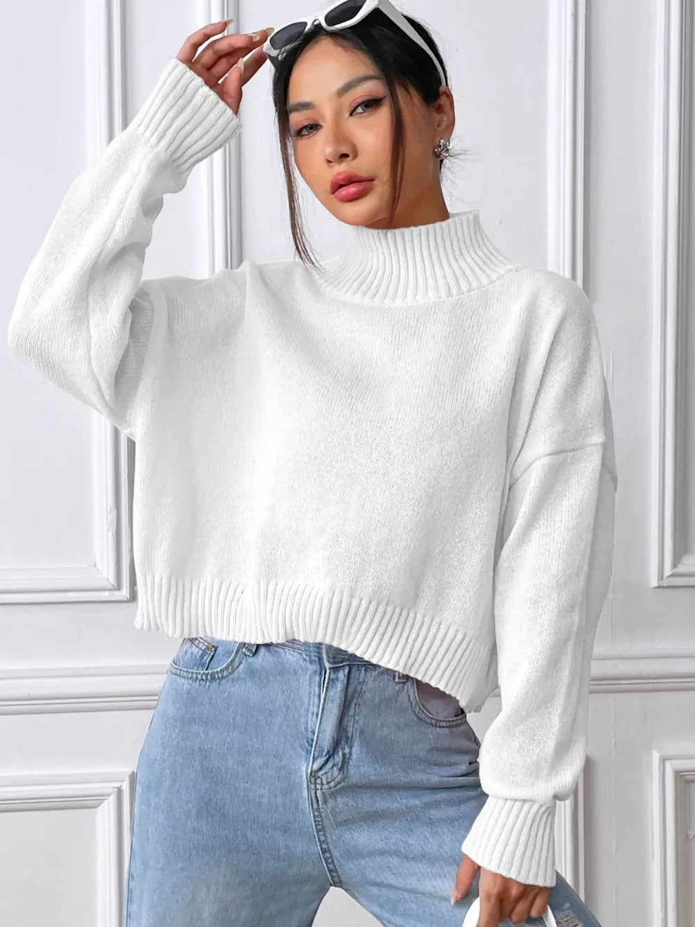 Person wearing a white sweater and blue jeans against a white paneled wall.