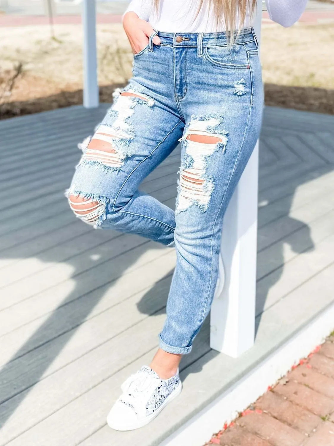 Person wearing distressed blue jeans and white sneakers on a wooden deck.