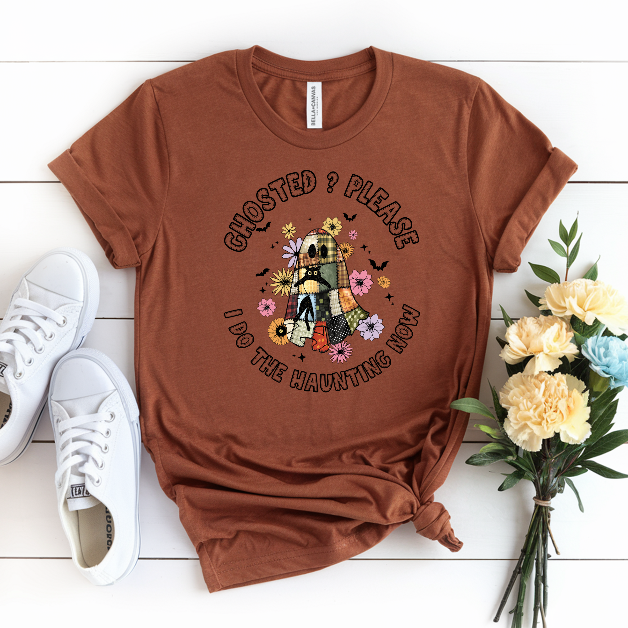 Brown t-shirt with ghost graphic and text on a white wooden surface with sneakers and flowers.
