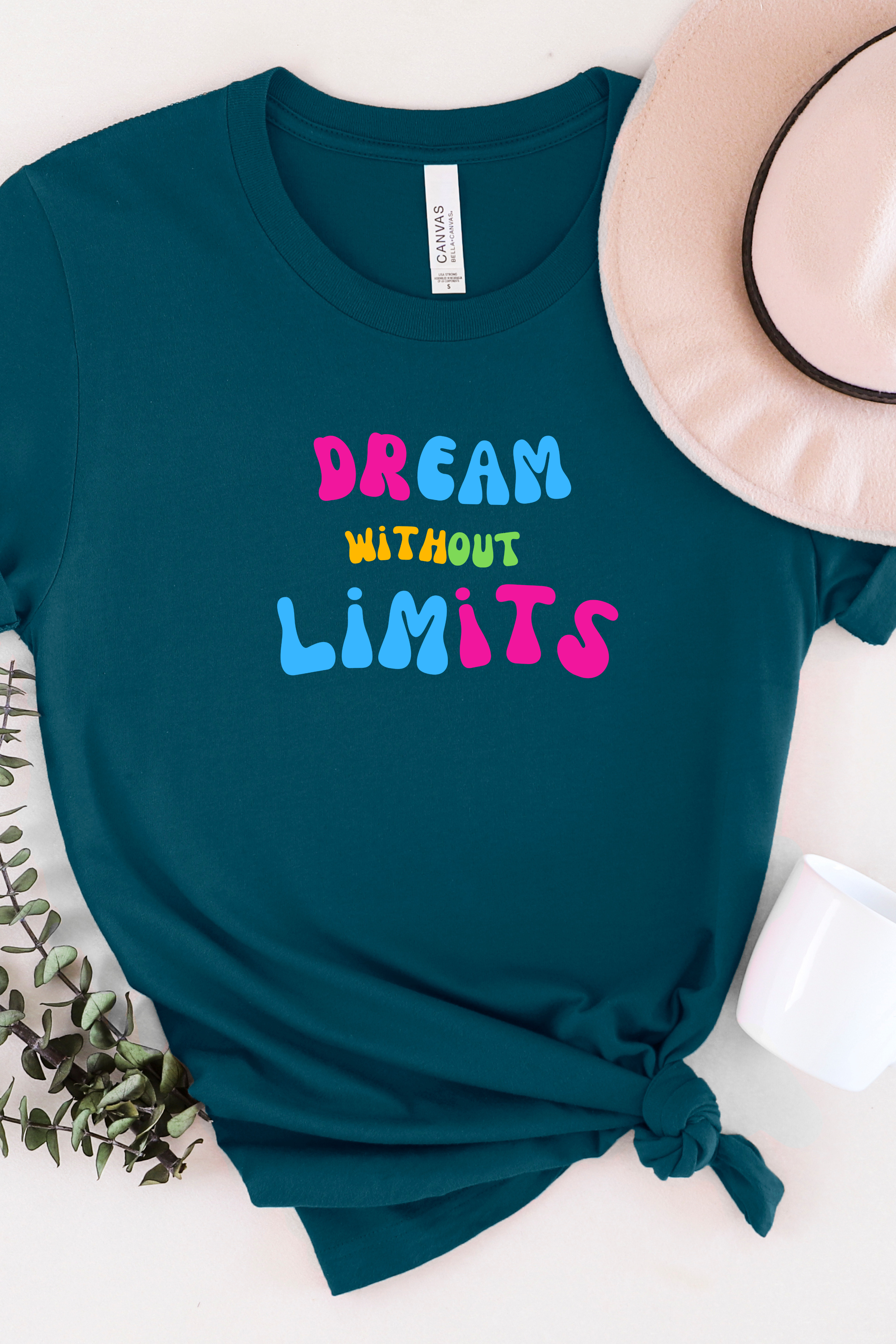 Teal t-shirt with 'Dream Without Limits' text on a white surface with a hat and plant.
